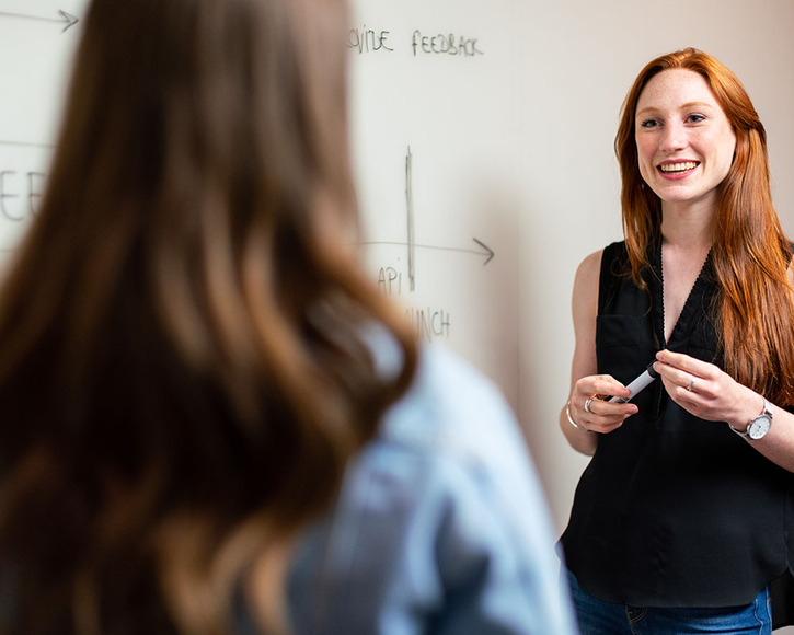 Young woman standing at a whiteboard addresses a female colleague