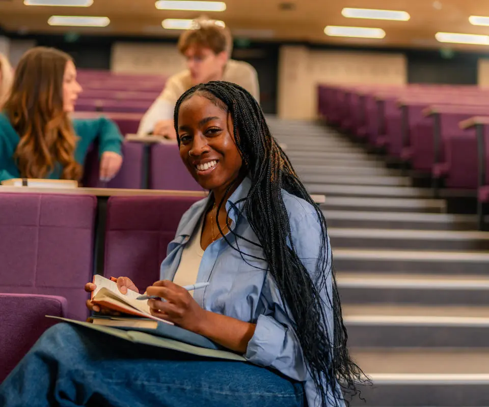 Female student in lecture theatre, making notes and smiling at camera