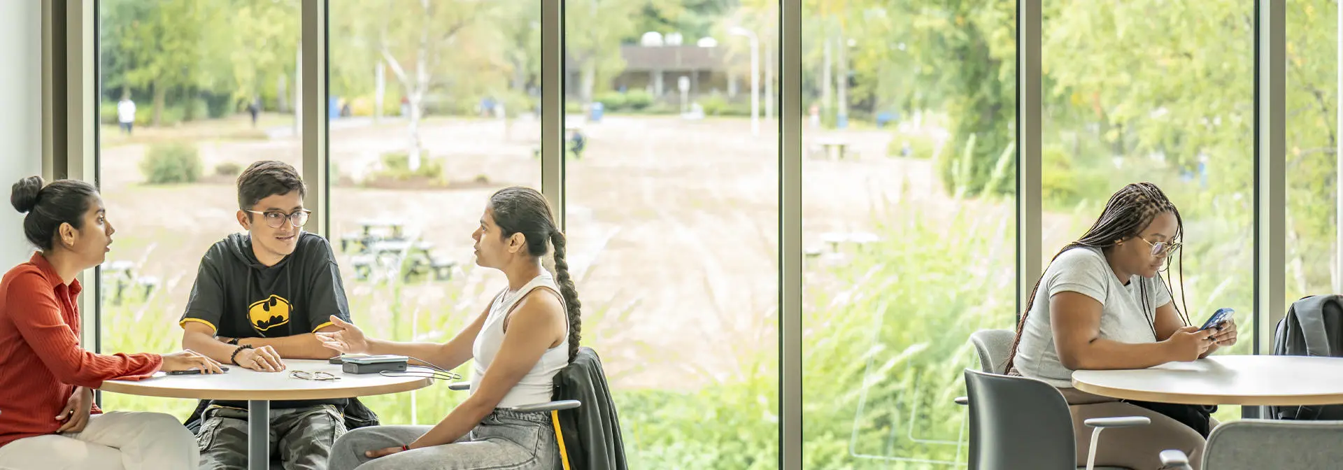 Four students sit at two round tables by large windows, three engaged in conversation and one focused on her phone.
