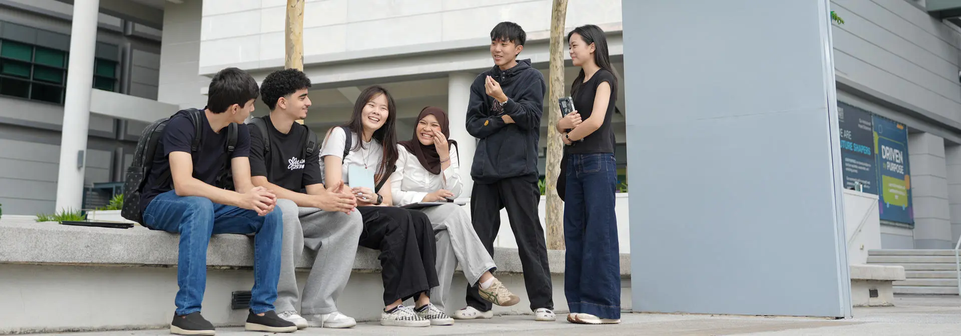A group of students sitting and standing on a bench outdoors, engaged in conversation.