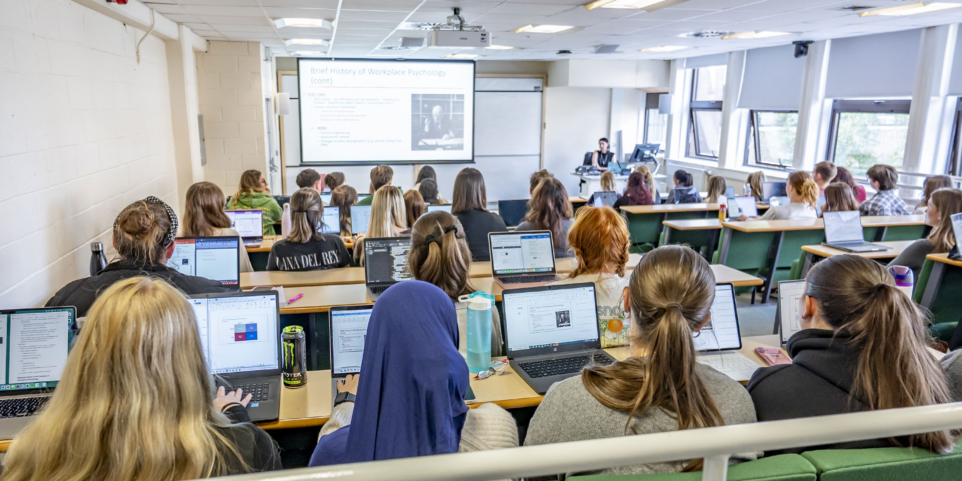 A classroom filled with students using laptops, with a projector displaying content at the front of the room.