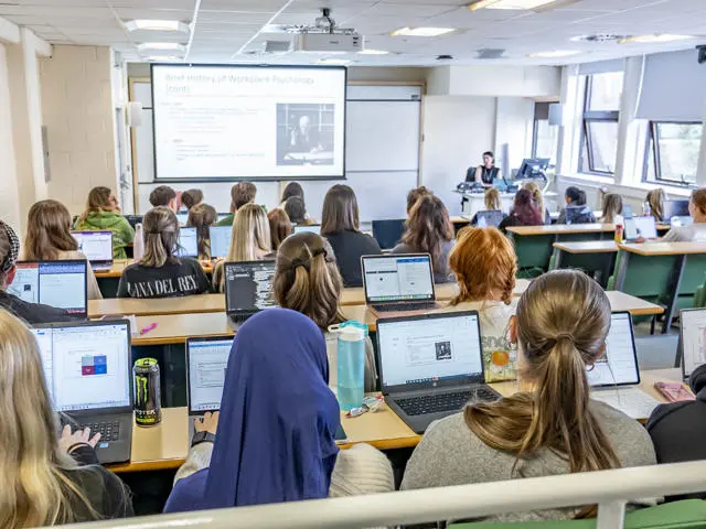 A classroom filled with students using laptops, with a projector displaying content at the front of the room.