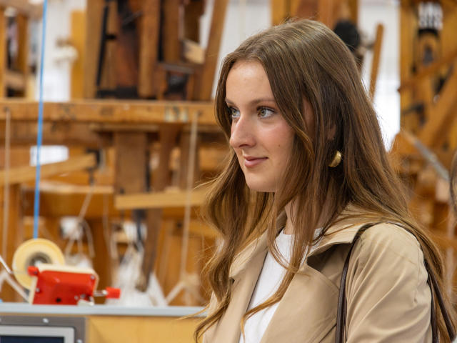 Young woman wearing a beige coat looking to the side, with a lot of wooden weaving machinery in the background.
