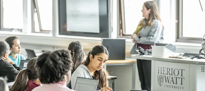 A woman teaches a class of students, standing in front of a screen displaying educational content.
