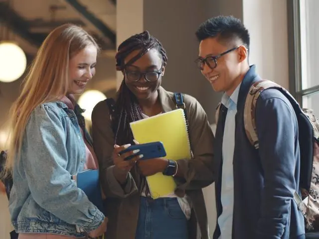 Three students standing in a corridor looking at a phone.