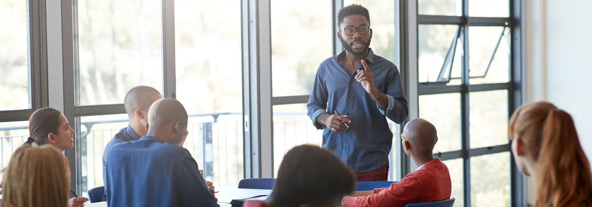 A man standing and leading a meeting consisting several people sitting down.