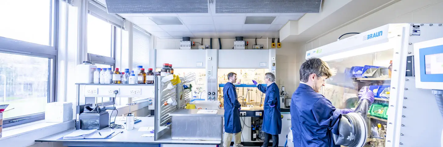 Two students in a chemistry lab, one using an isolation glove box and one talking with a member of staff