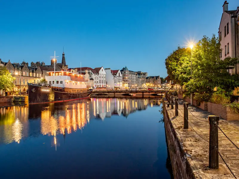 A city river at dusk, featuring a boat on the water, surrounded by evening lights.
