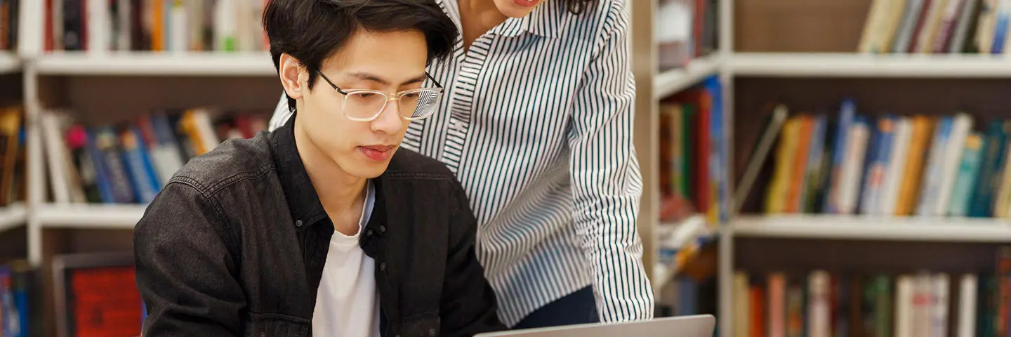 A man works on a laptop in a library with support of another behind him.