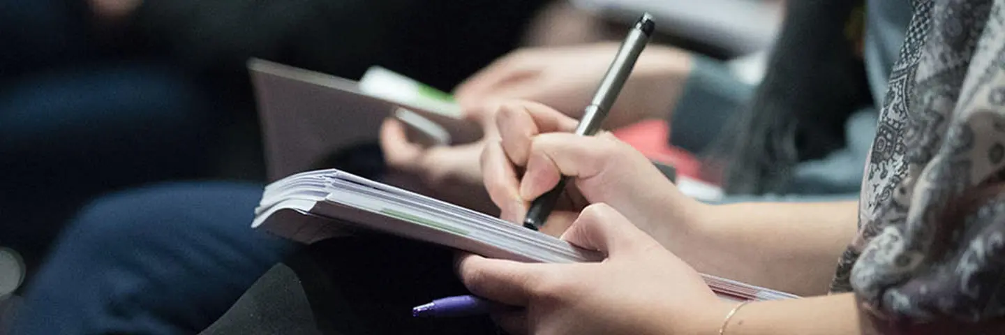 Person sitting at an event taking notes with a pen on a notepad.