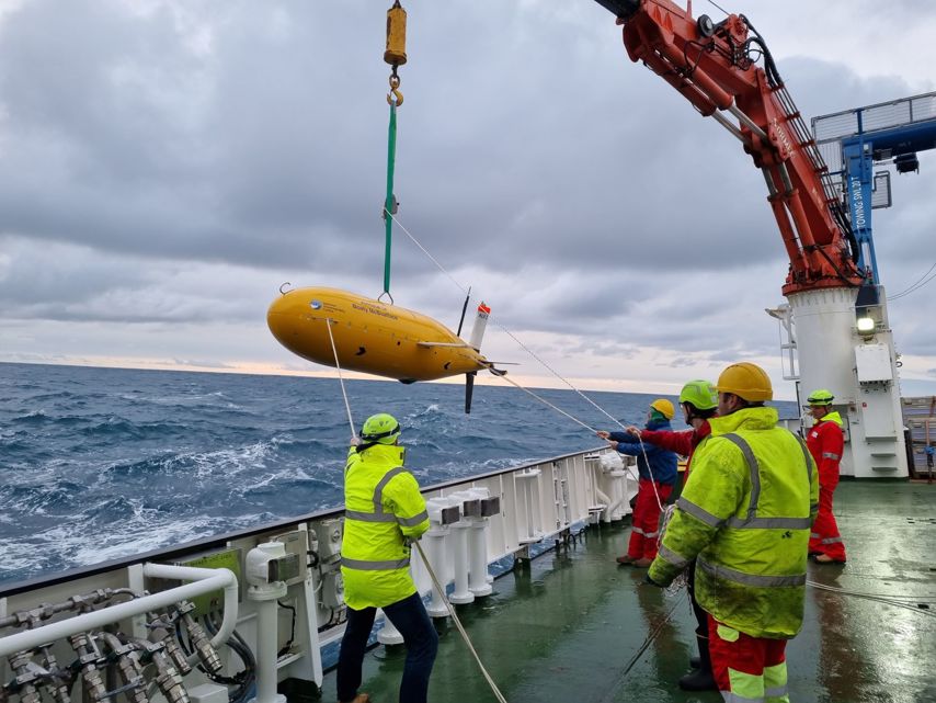Researchers worked with autonomous vehicle, including Boaty McBoatFace The image shows a group of individuals on the deck of a ship, wearing high-visibility clothing and hard hats. They are engaged in deploying or retrieving a large yellow torpedo-shaped object using a crane.