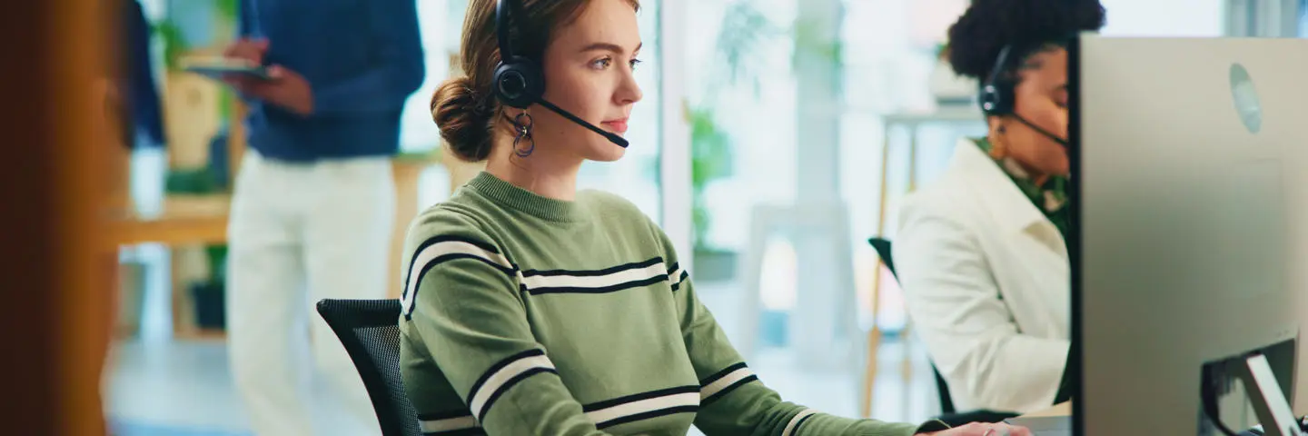 A woman wearing a headset sits at a computer on the phone.