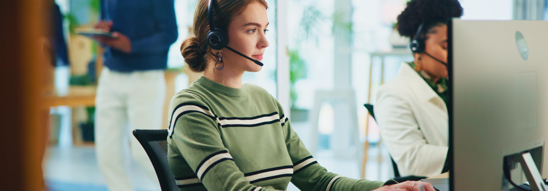 A woman wearing a headset sits at a computer on the phone.