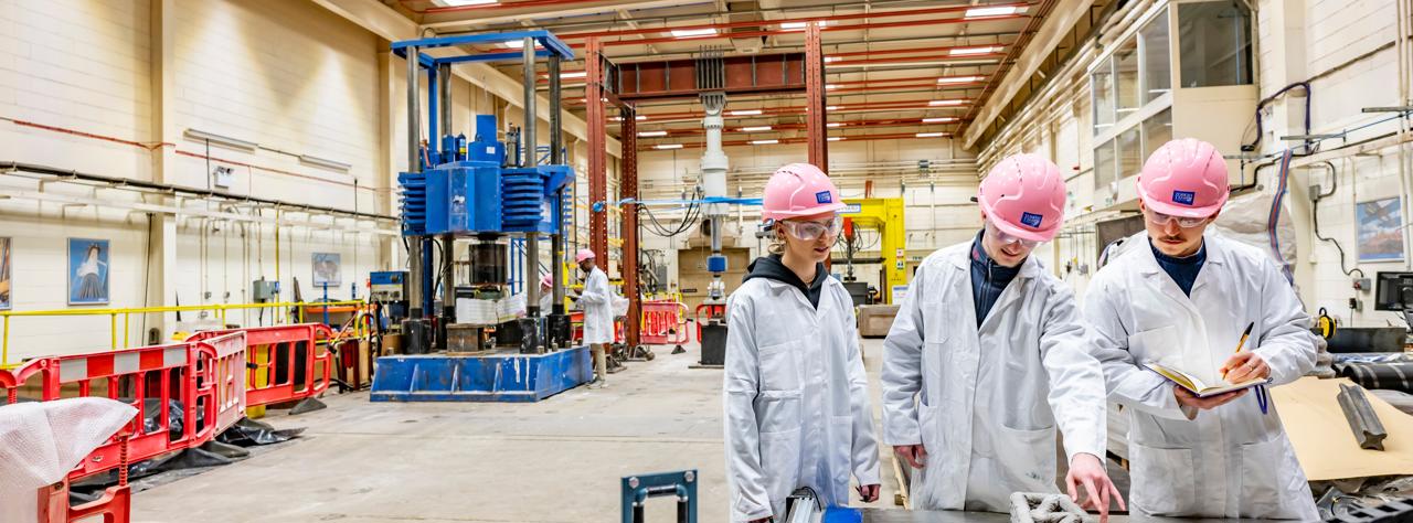 Three individuals wearing lab coats and safety helmets in warehouse.