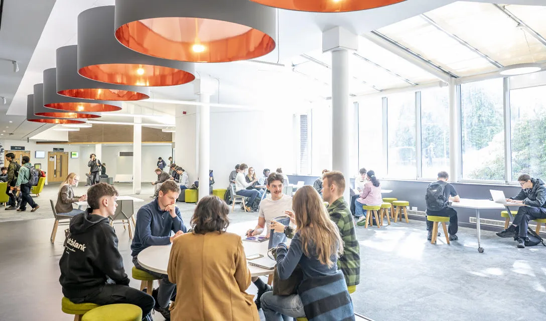Students collaborate at a table in a spacious, open area filled with natural light and modern furnishings.