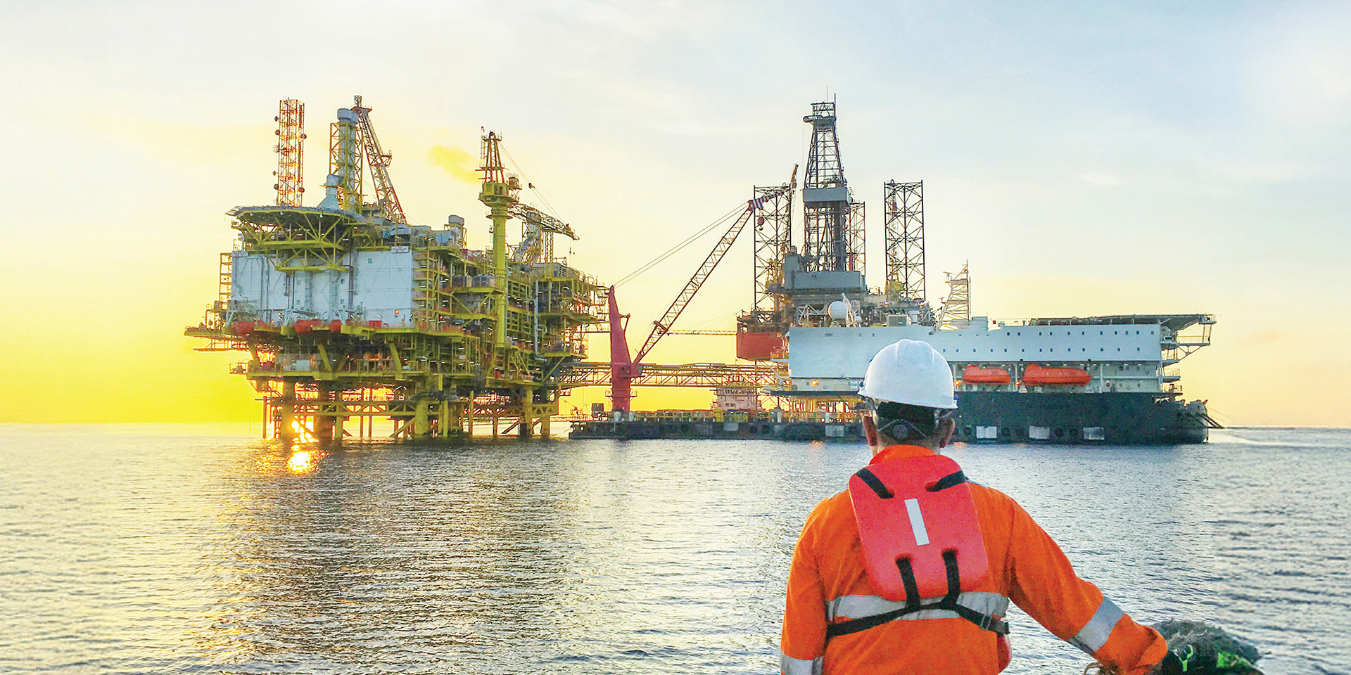 A man dressed in an orange safety vest stands before an oil rig, showcasing safety protocols in a construction setting.