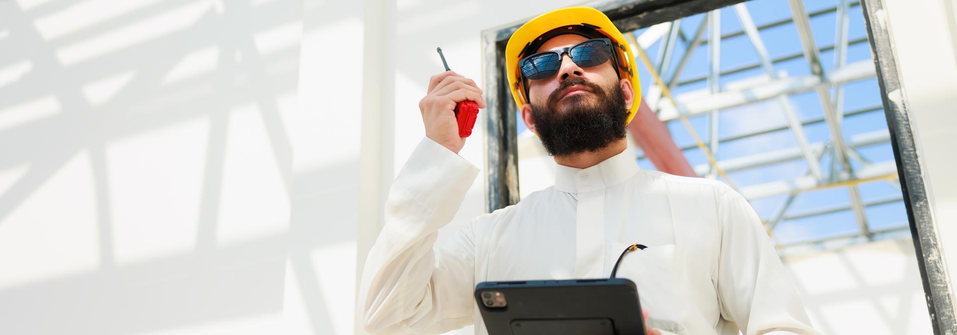 civil engineer in kandura and yellow hard hat inspecting a site talking into a walkie-talkie