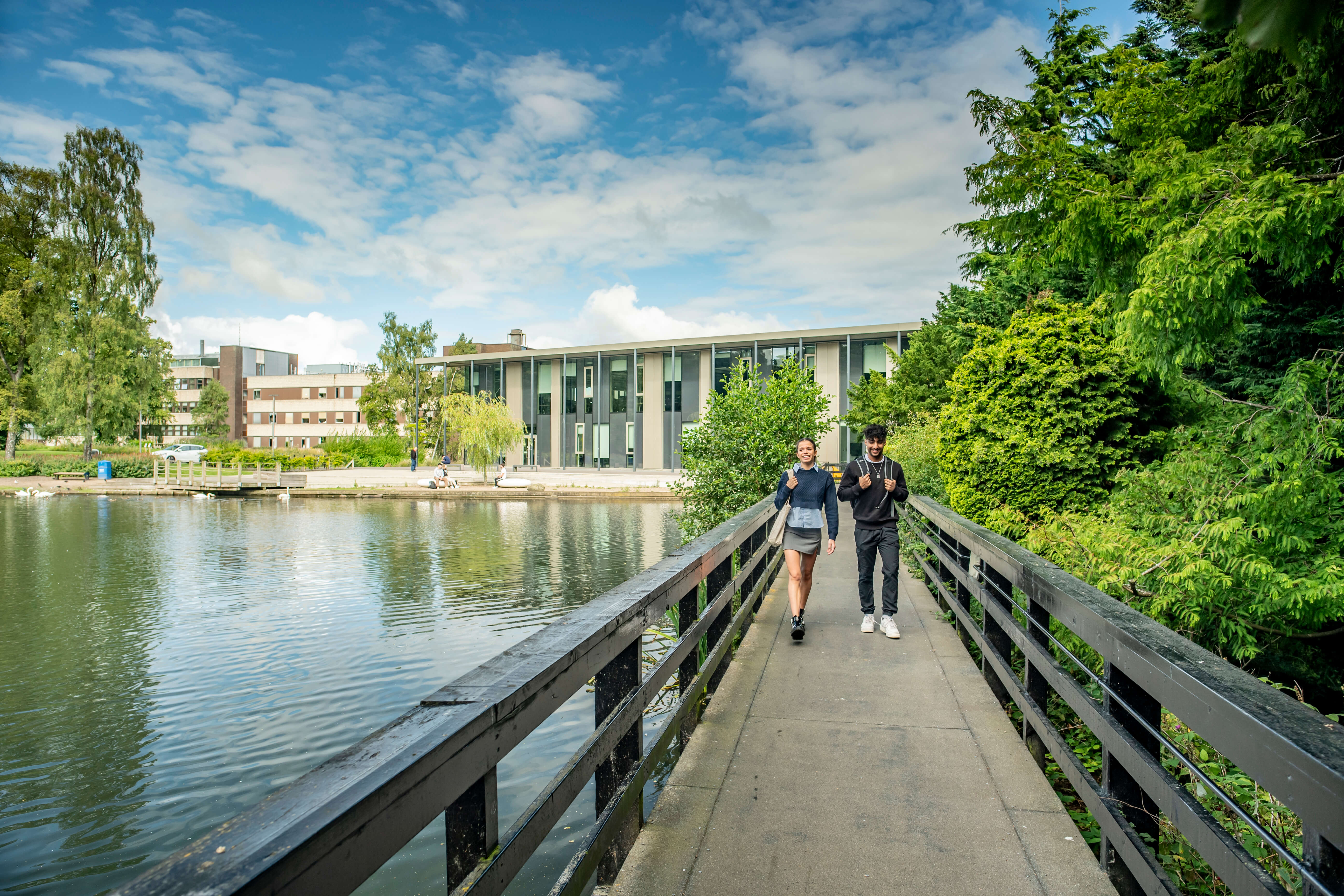 Two students walking on a bridge over a loch, with trees on one side and a modern building in the distance