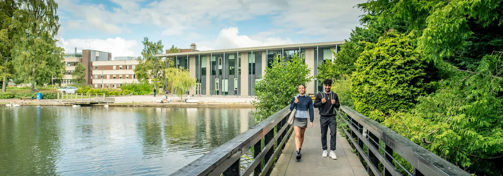 Two students walking on a bridge over a loch, with trees on one side and a modern building in the distance