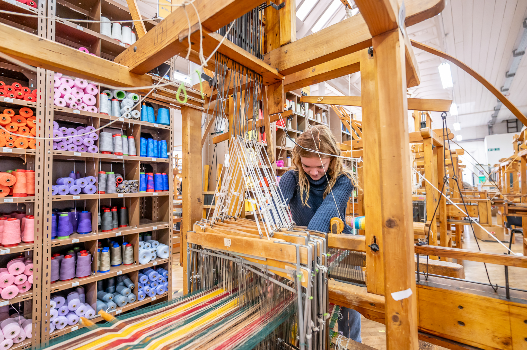 A woman works with threads and materials on a wooden weaving machine.