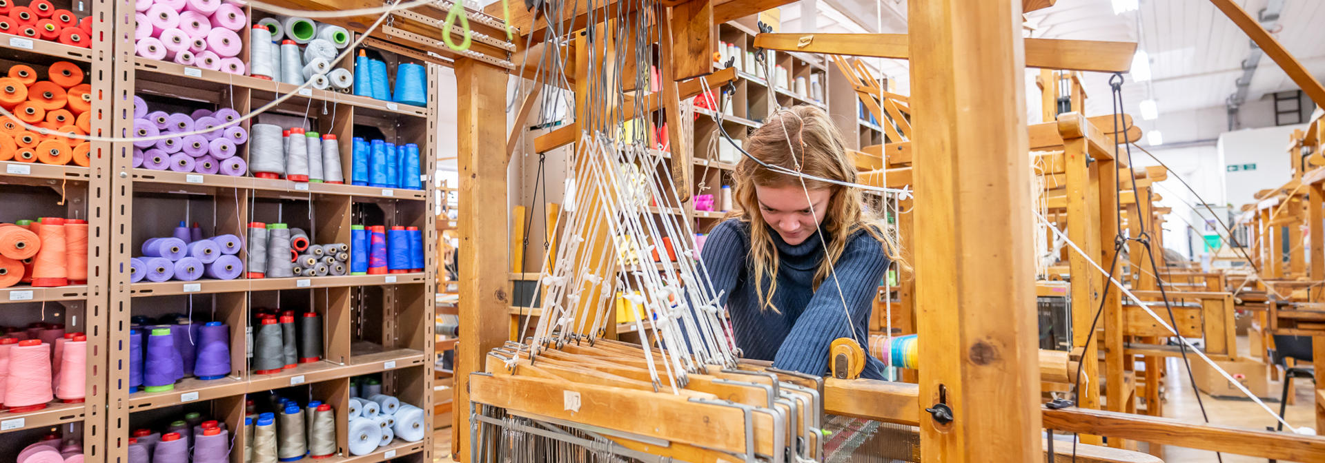 A woman works with threads and materials on a wooden weaving machine.