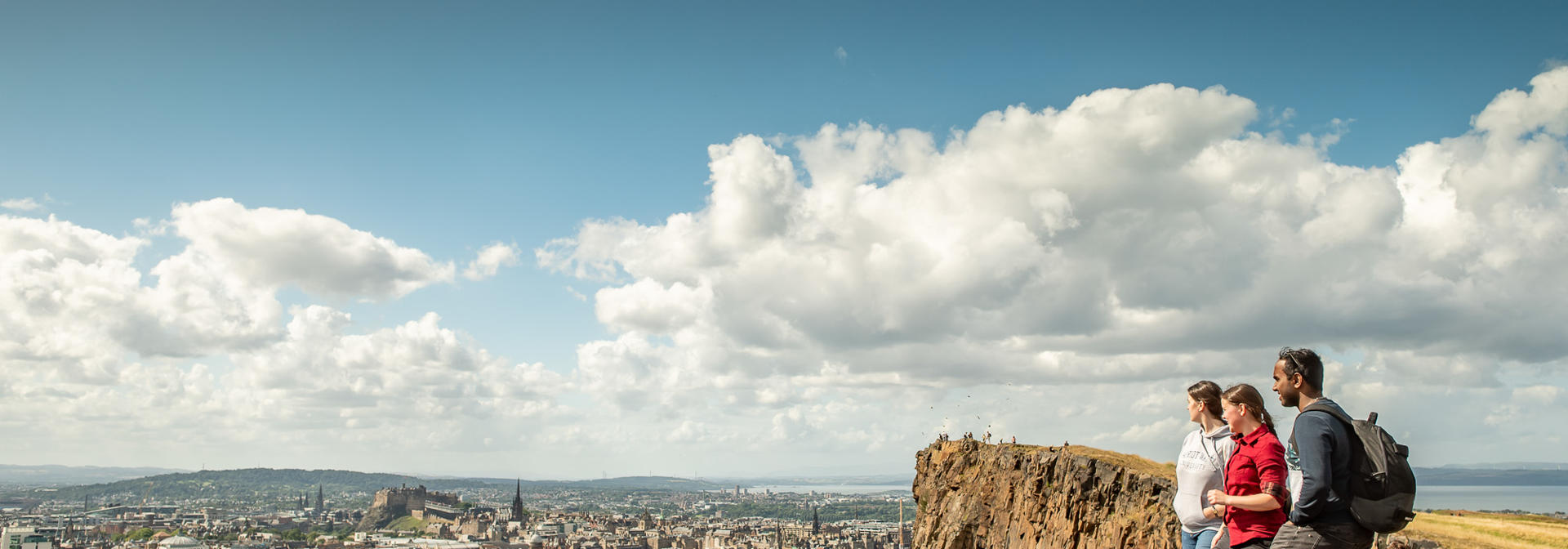 Three individuals stand atop a hill and look over at a city below.
