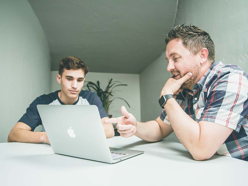 A man points out something on his laptop screen to a younger man