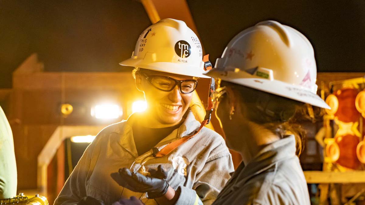 Two women marine engineers in overalls and safety helmets discuss a mollusc taken from the sea