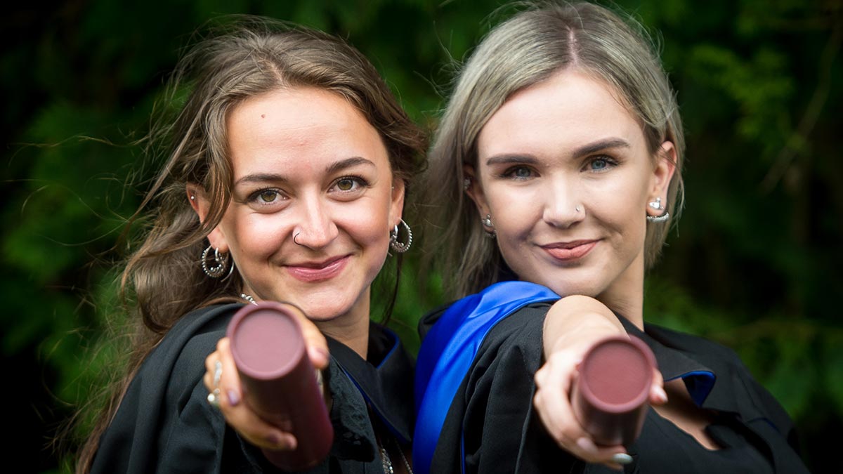 Two female graduates in robes point their scrolls at the camera